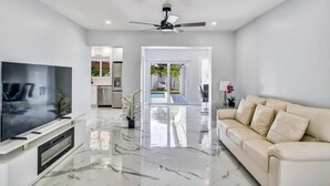 A modern living room with a flat-screen TV, a beige sofa, and a ceiling fan. - TropiCasa - new construction (Oakland Park)