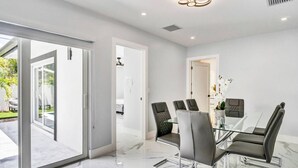 A modern dining area with a glass table and metal chairs, a white interior, and a view of the outdoors. - TropiCasa - new construction (Oakland Park)