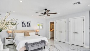A modern bedroom with a large bed, bedside tables, a ceiling fan, and a bathroom visible through an open door. - TropiCasa - new construction (Oakland Park)