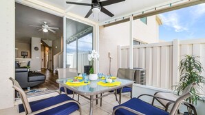 Un patio con una mesa de vidrio, sillas con cojines azules, un ventilador de techo y una planta en maceta.