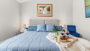 Un dormitorio con una cama, una mesita de noche, una silla azul, una bandeja con comida y bebidas, y un cuadro en la pared.