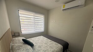 Quarto pequeno e bem organizado com uma cama de casal, uma janela com persianas, um ar condicionado e uma pequena mesa de cabeceira.