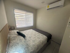 Quarto pequeno e bem organizado com uma cama de casal, uma janela com persianas, um ar condicionado e uma pequena mesa de cabeceira.