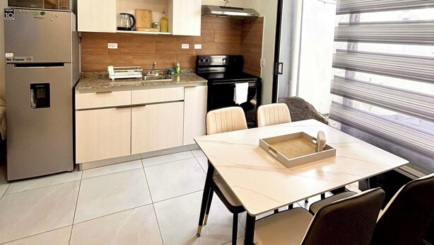 A compact kitchen with a refrigerator, stove, and sink. There is a dining table with chairs and a window with blinds.