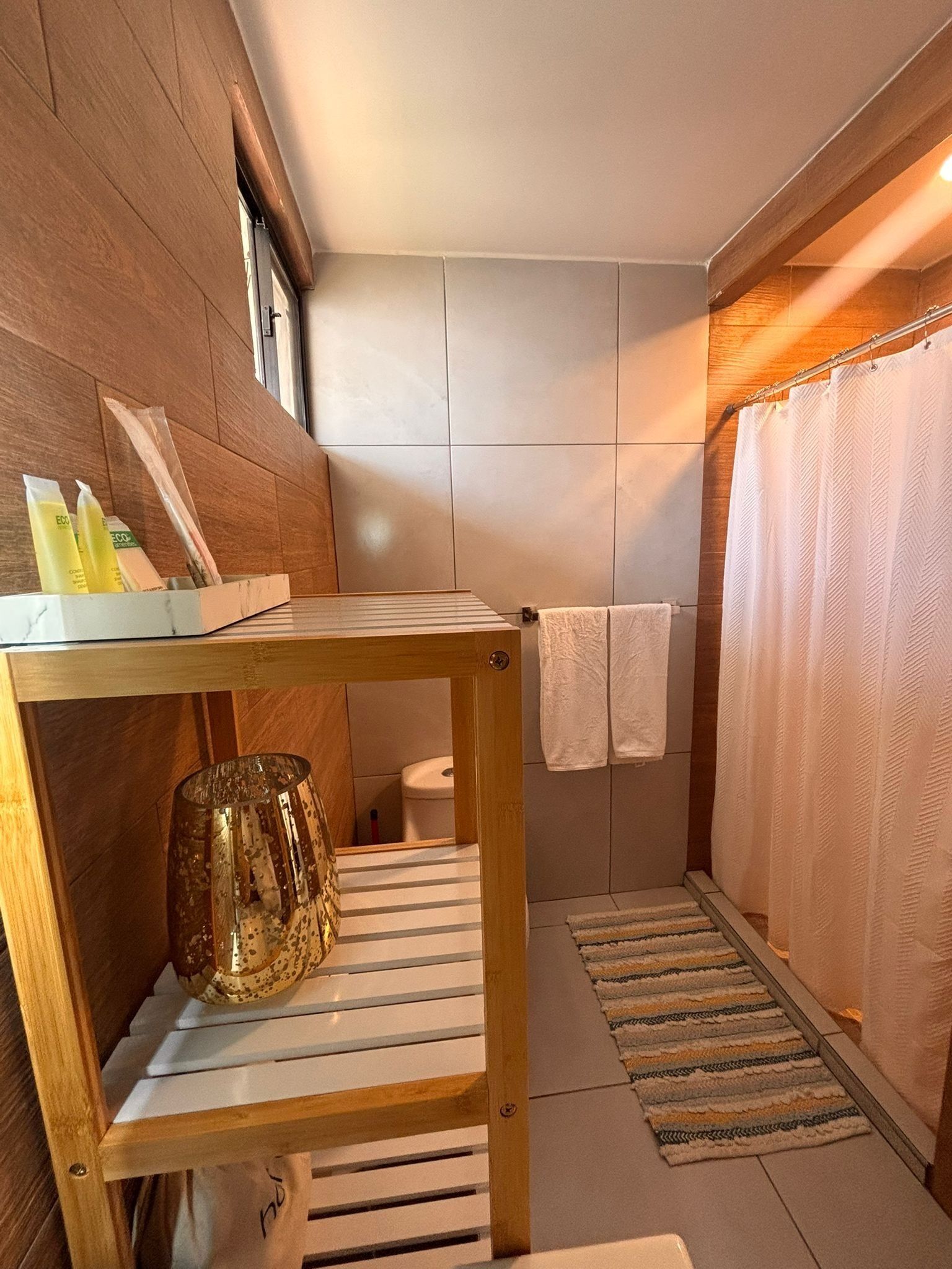 A bathroom with a wooden vanity, a glass vase, a striped towel, a shower curtain, and a small rug.