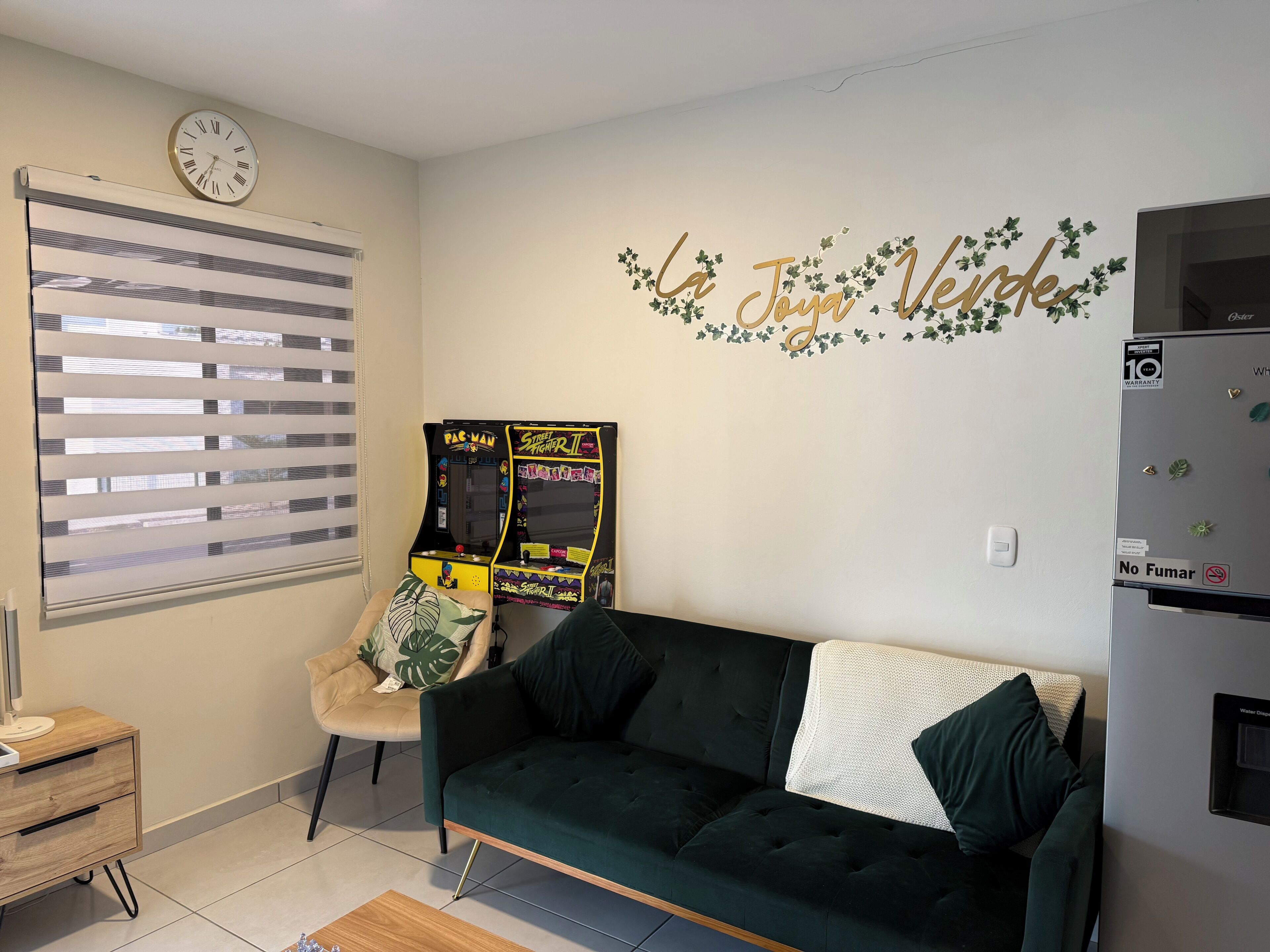 Una sala de estar con un sofá verde oscuro, un sillón color beige, un reloj y dos máquinas de videojuegos.