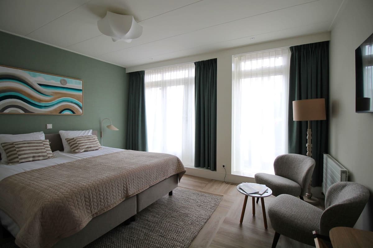 A hotel room with a bed, two armchairs, a small table, a lamp, and a painting on the wall.