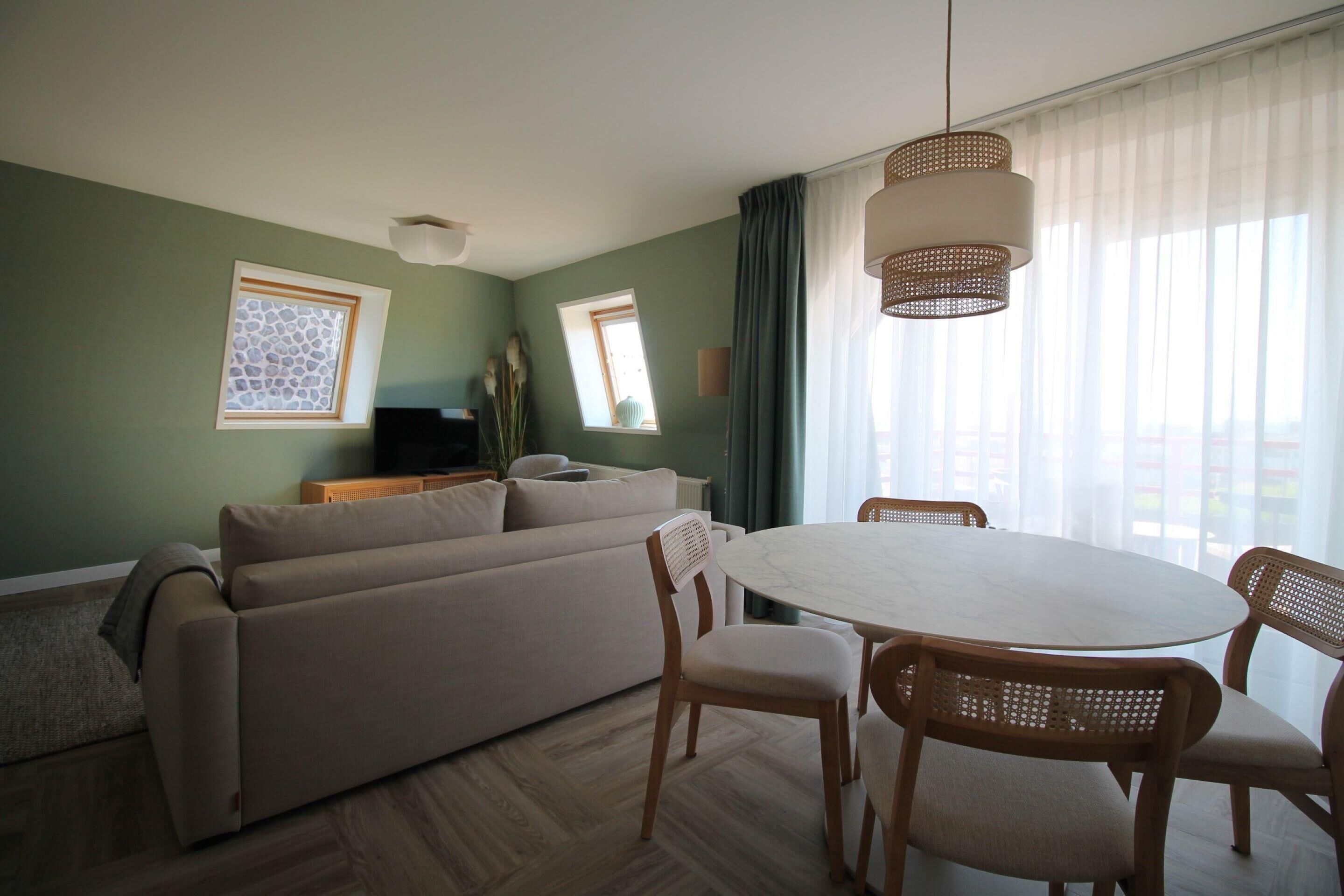 Un salon moderne avec un canapé, une table à manger et des chaises. Il y a deux fenêtres avec des rideaux, un téléviseur posé sur un meuble et un luminaire suspendu.