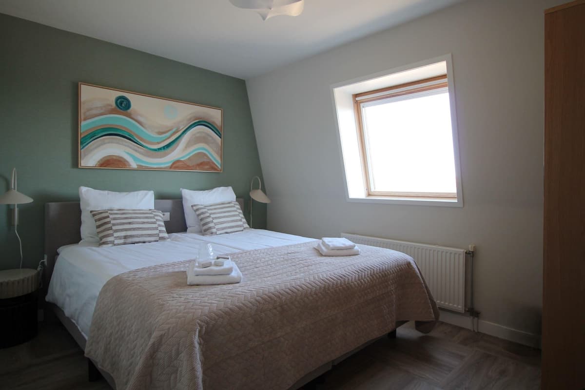 A bedroom with a bed, pillows, a painting, a window, and a radiator.