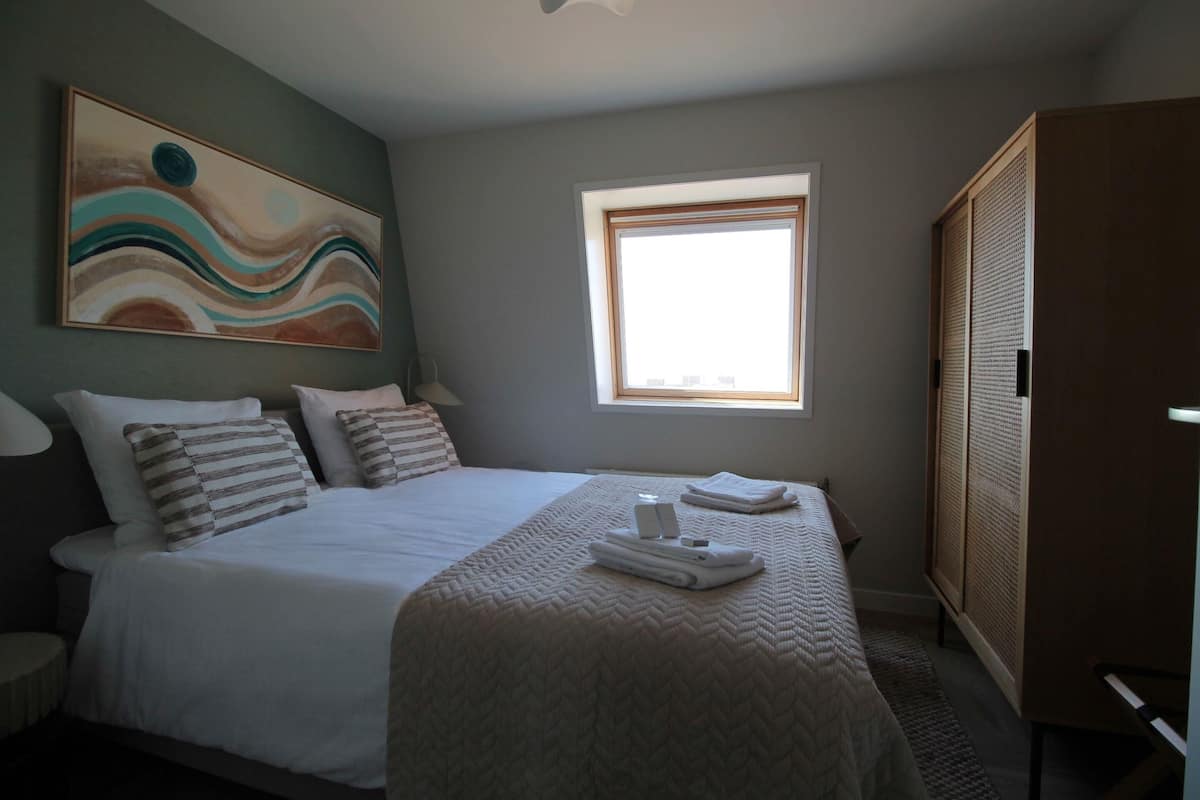 A hotel room with a bed, a window, a painting on the wall, and a wardrobe.