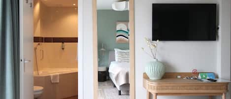 A hotel bathroom with a shower, toilet, and a mirror reflecting a bed and a wall-mounted TV.
