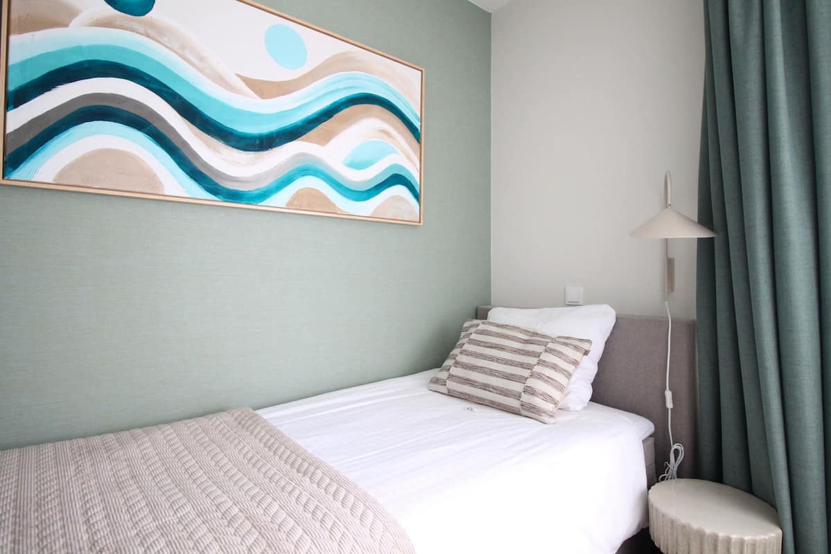 A neatly made bed with a striped pillow, a wall-mounted abstract painting, a bedside lamp, and a curtain.