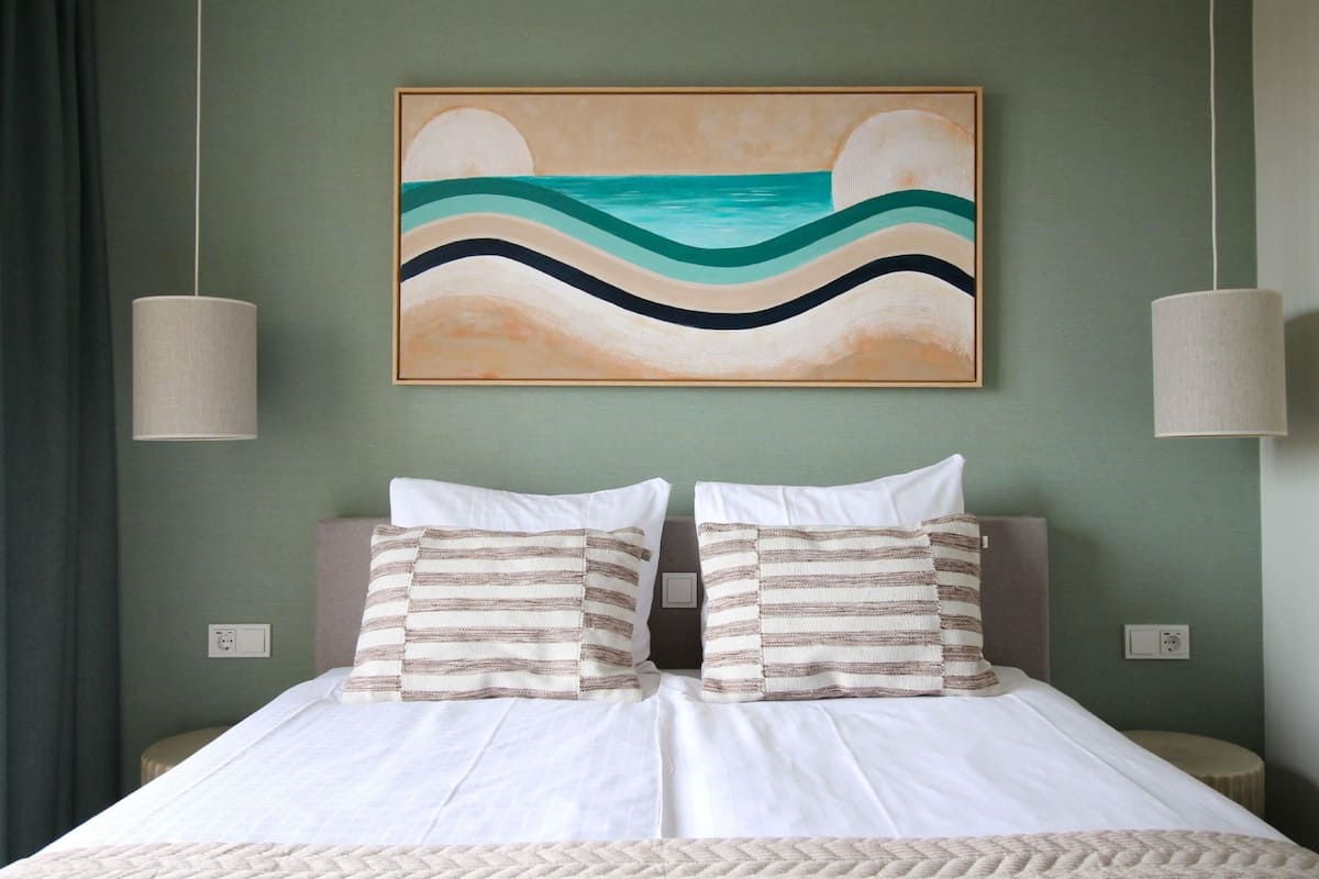 A neatly made bed with white linens and striped pillows, a painting of a beach scene above it, and two pendant lights hanging from the ceiling.