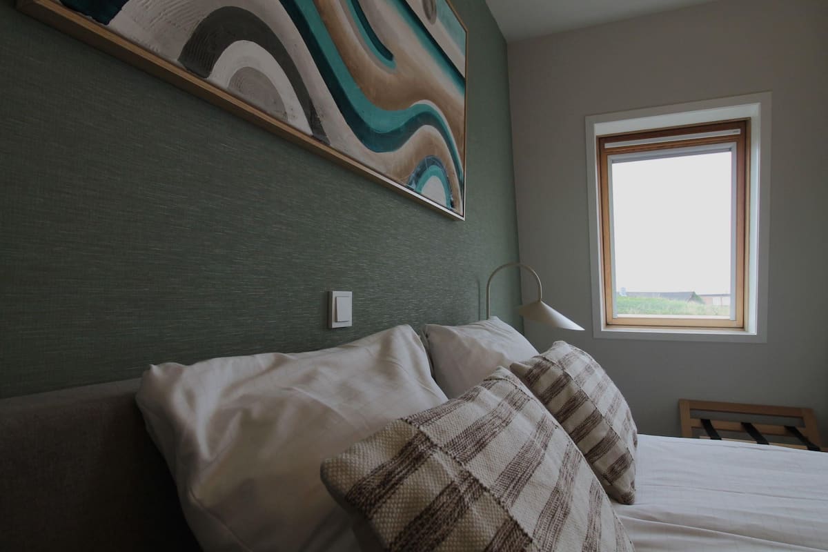 A neatly made bed with white linens and plaid pillows, a modern abstract painting on the wall, a bedside lamp, and a window with a view of greenery.