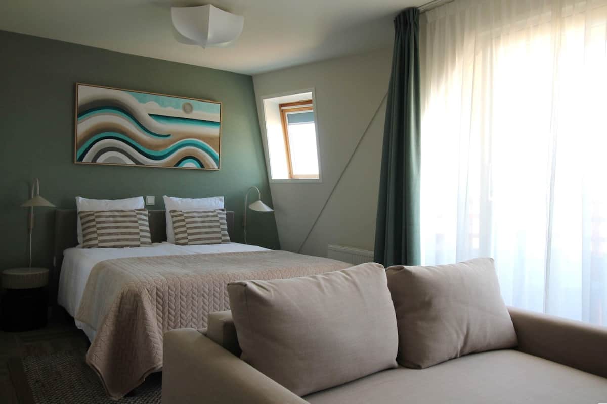 A hotel room with a bed, a sofa, a painting, a window, and a lamp.