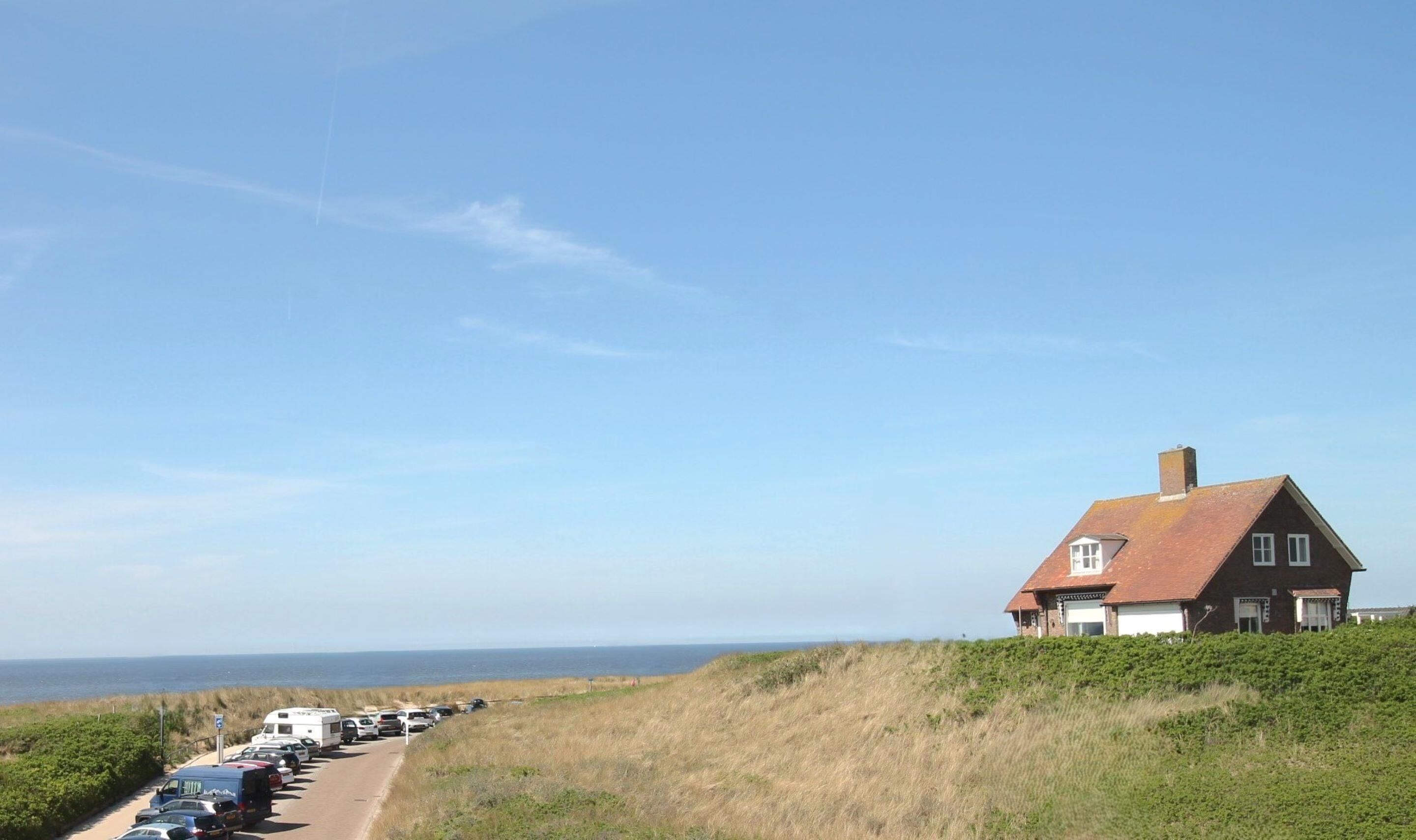 A coastal house with a long driveway lined with parked vehicles, overlooking the sea.