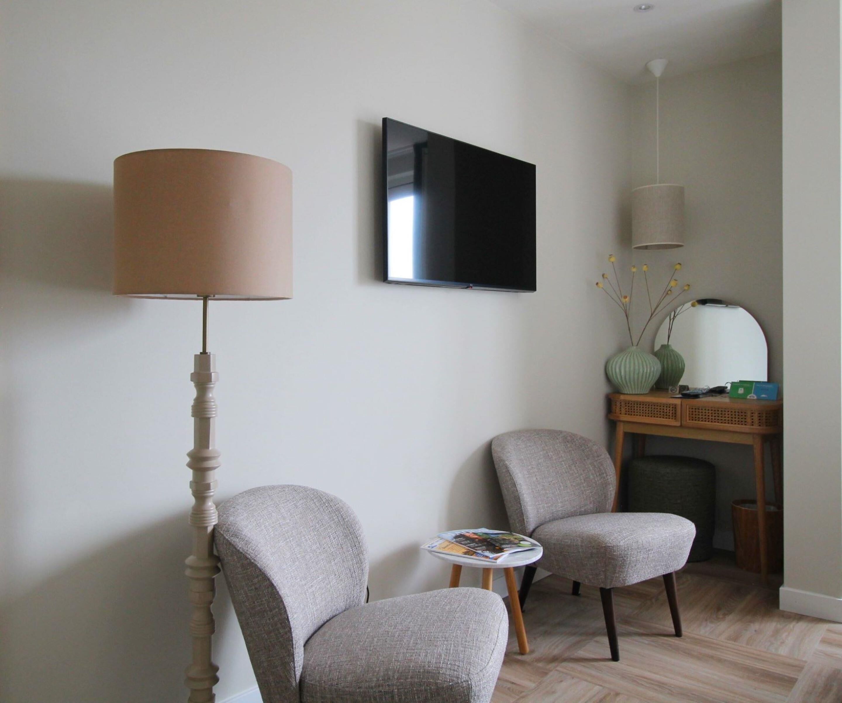 A modern living room with two grey armchairs, a round table, a flat-screen TV, a lamp, and a small side table with decorative items.