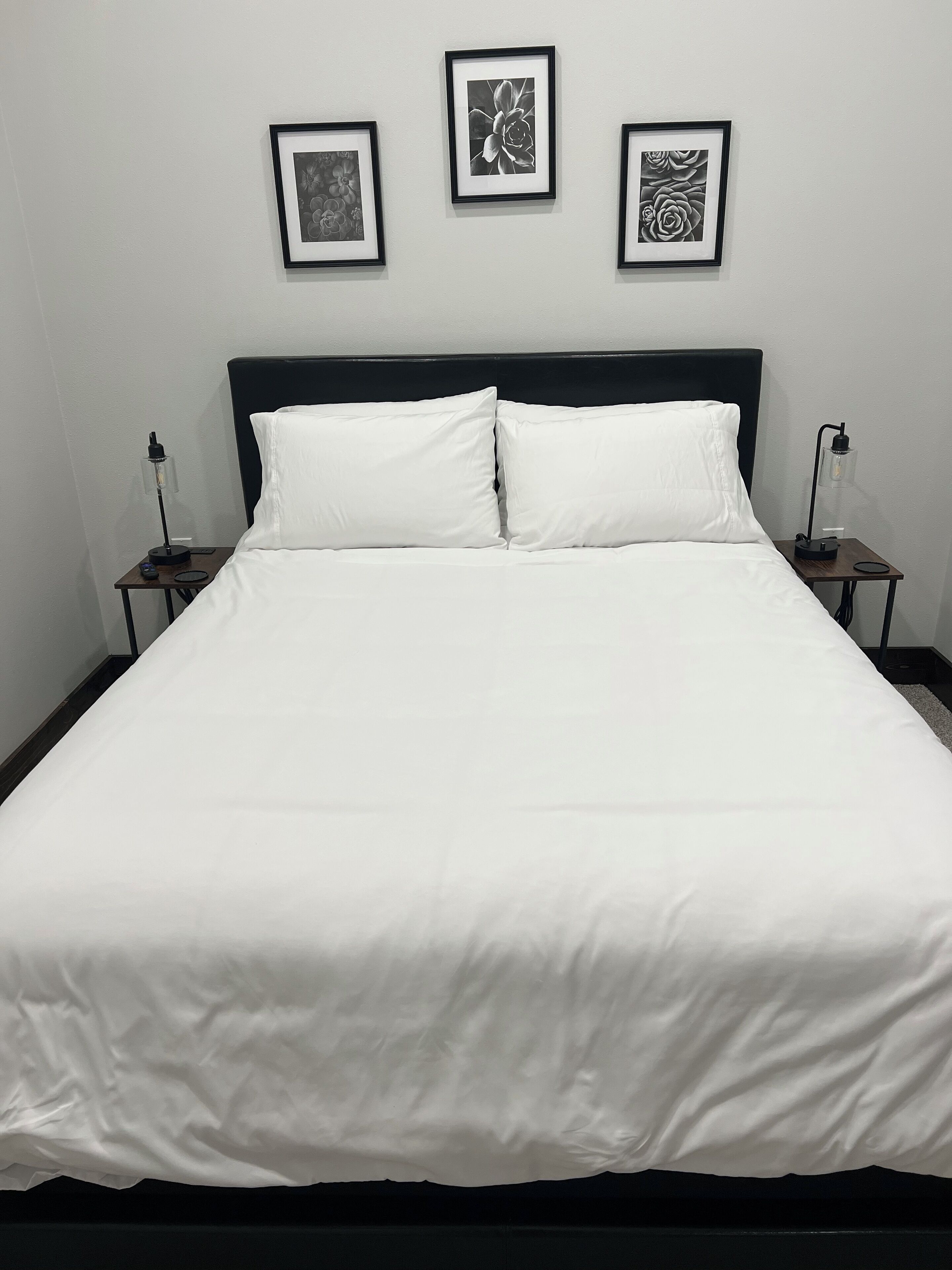 Un lit bien fait, avec une literie blanche, deux tables de chevet avec des lampes et trois œuvres d’art encadrées accrochées au mur.