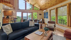 A spacious living area with a dark sofa, a wooden coffee table, and a dining table with chairs. A ceiling fan is visible, and large windows offer a view of the outdoors.