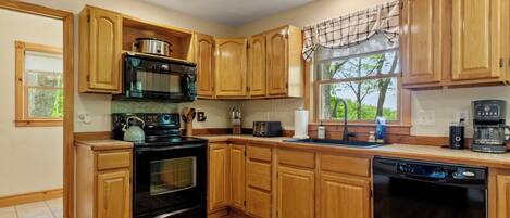 A kitchen with wooden cabinets, a stove, a microwave, a dishwasher, and a window with a view of trees.