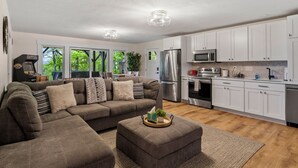 A modern living room with a sectional sofa, a dining area with a table and chairs, and a kitchen with white cabinets and stainless steel appliances.