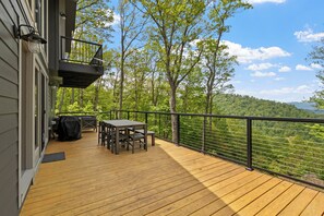 Eine Holzterrasse mit Tisch und Stühlen, einem Grill und Blick auf einen bewaldeten Hang.