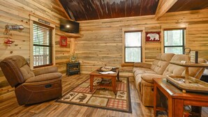 L'intérieur d'une cabane en rondins, avec un fauteuil inclinable en cuir, un canapé, une table basse et un téléviseur fixé au mur.