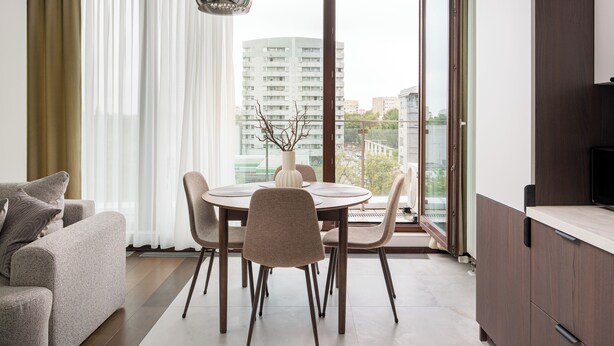 Uma sala de estar moderna com sofá, mesa de jantar e cadeiras, uma janela grande com cortinas e vista para edifícios.
