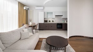 Uma sala de estar moderna com um sofá, uma mesa de centro e uma área de jantar com mesa e cadeiras. Uma cozinha visível ao fundo, com armários e eletrodomésticos.