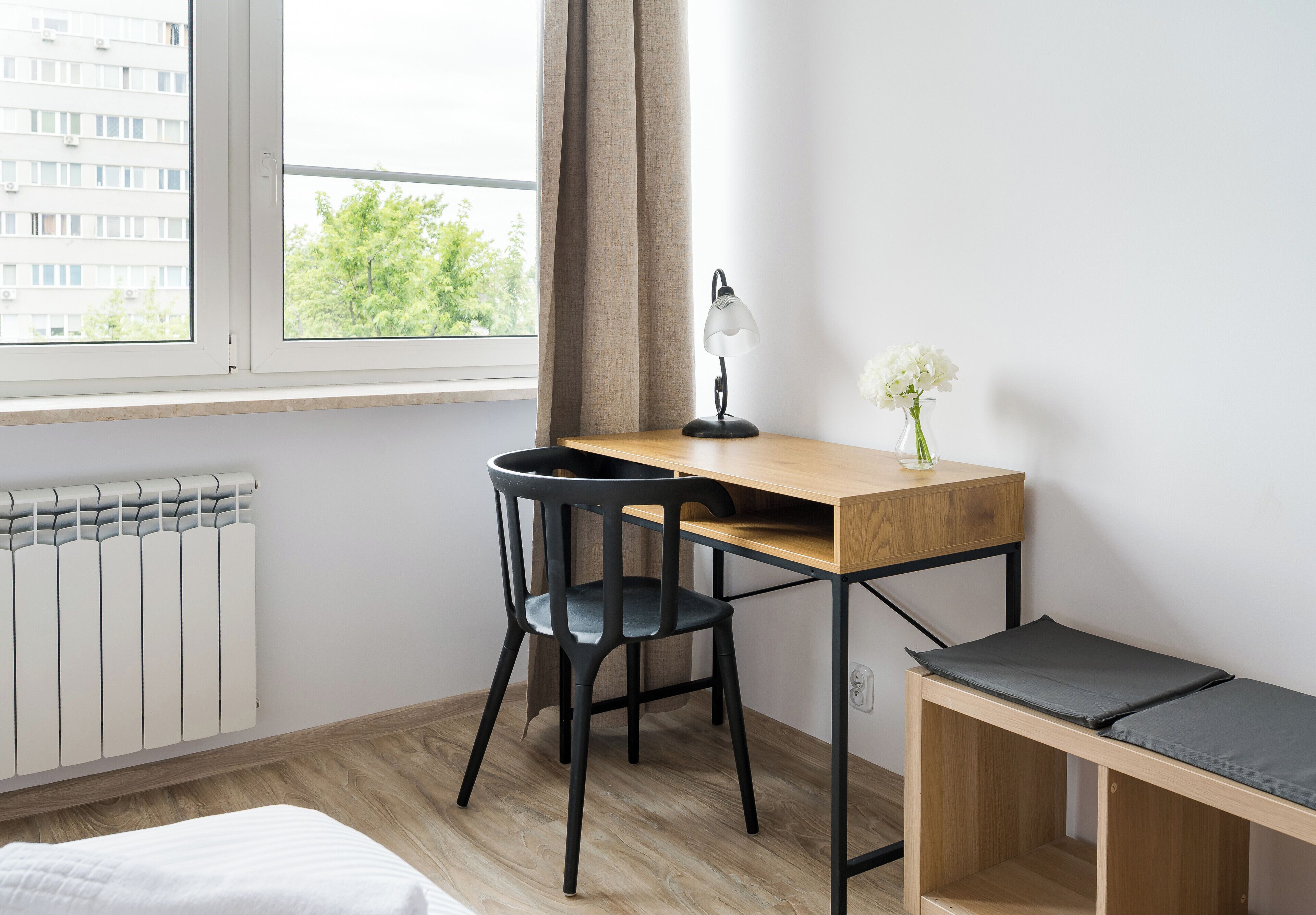 Ein modernes Schlafzimmer mit einem Holztisch, einem schwarzen Stuhl, einem Nachttisch mit Kissen, einer Heizung und einem Fenster mit Blick auf Gebäude und Bäume.