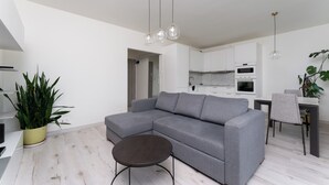 A modern living room with a grey sectional sofa, a small round table, and hanging pendant lights. There is a potted plant and a dining area with a table and chairs.