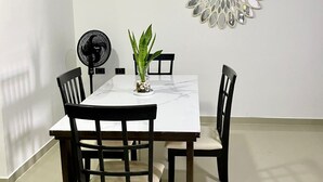 Et spiseområde med et hvidt bord, sorte stole, en potteplante, et spejl på væggen og en gulvventilator.