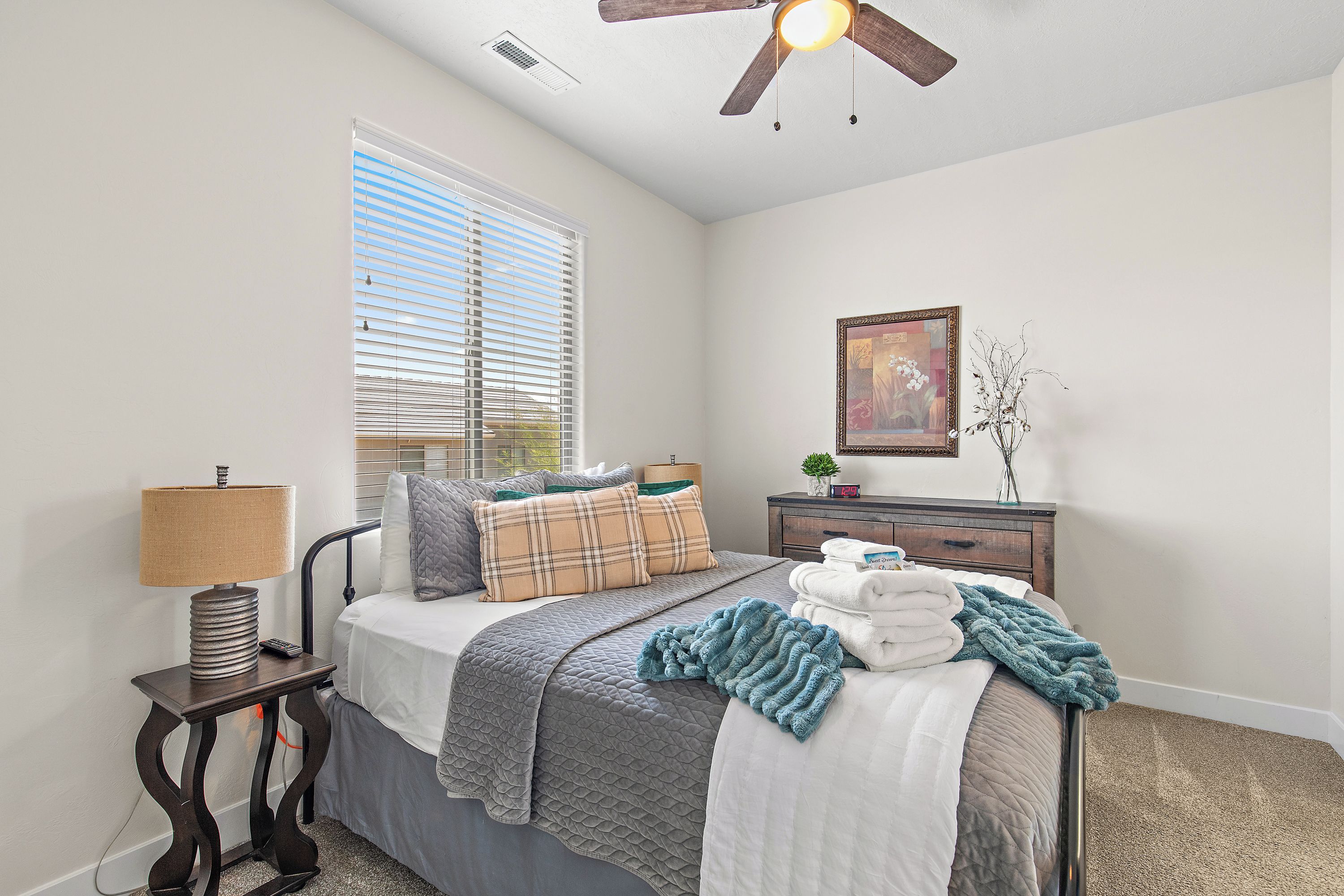 A bedroom with a bed, bedside table, lamp, window with blinds, ceiling fan, dresser, and a framed picture on the wall.