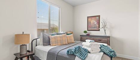 A bedroom with a bed, bedside table, lamp, window with blinds, ceiling fan, dresser, and a framed picture on the wall.