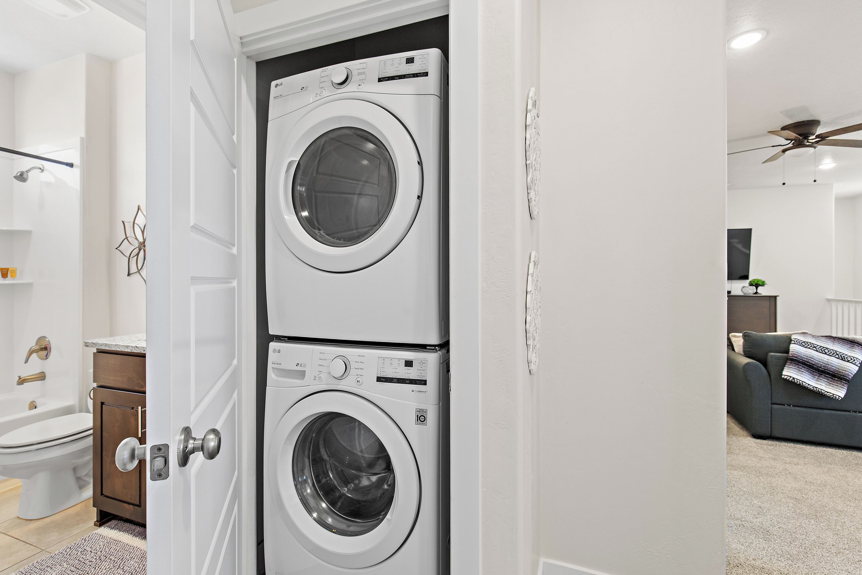 A laundry room with two stacked front-loading washing machines, a door with a silver handle, and a ceiling fan in the adjacent living area.