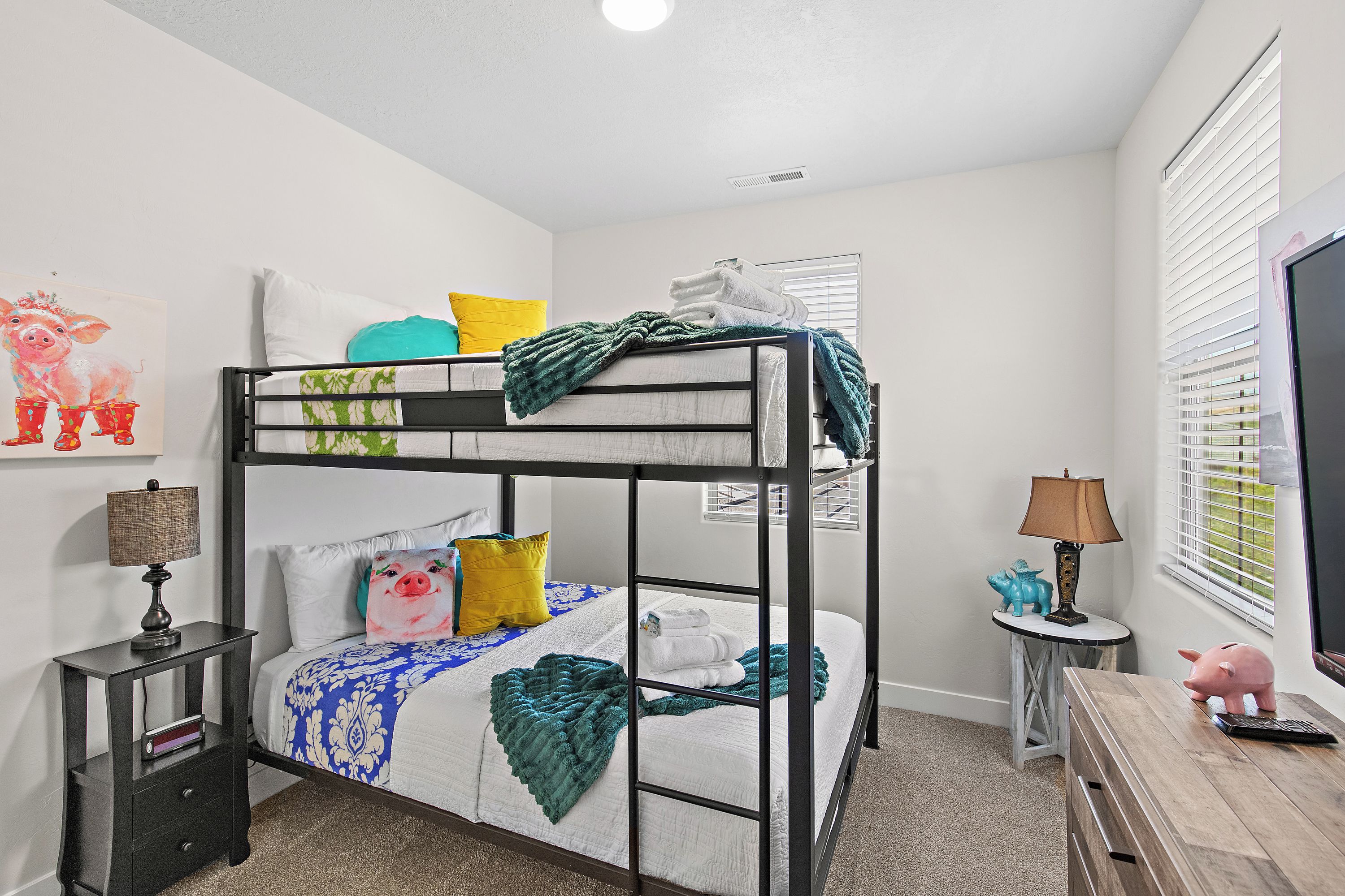 A bunk bed with a white mattress, teal and yellow pillows, and a green blanket. There is a small table with a lamp and a piggy bank.