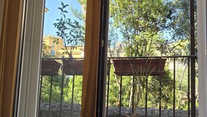 Een kamer met een schuifdeur naar een buitenbalkon, met een bloempot en uitzicht op bomen en een gebouw.
