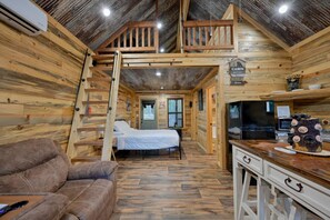 A cabin interior with a loft bedroom, a sofa, a kitchen area, and a wooden exterior.