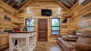 A log cabin interior with a wooden dining table, a sofa, and a television mounted on the wall. - Cozy Cabin 3 miles to WPG Slps 4, ATV friendly (Mena)
