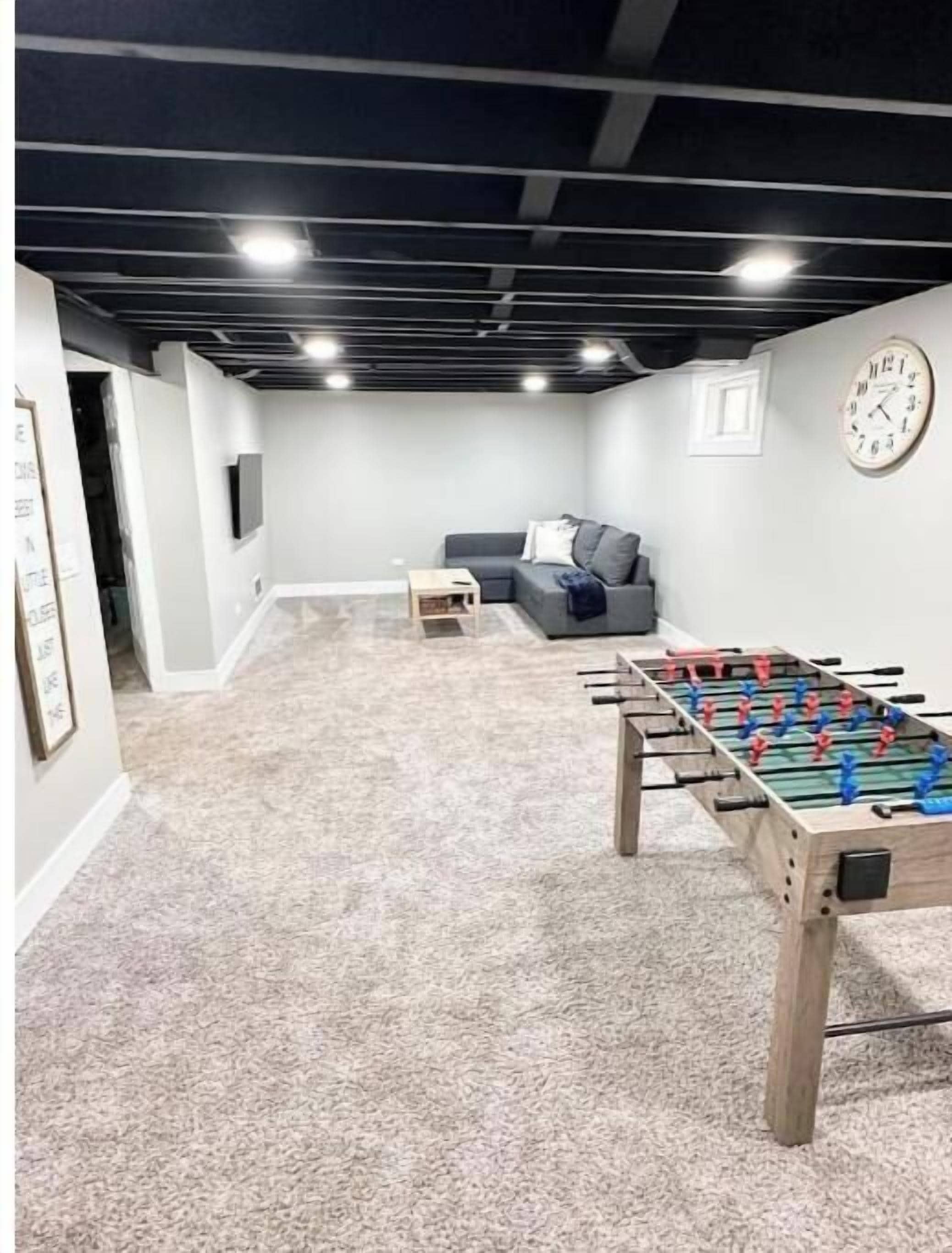 A room with a foosball table, a grey sofa, a small table, and a clock on the wall.