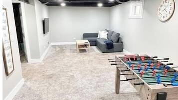 A room with a foosball table, a grey sofa, a small table, and a clock on the wall.
