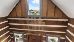 Une chambre de style rustique, avec un plafond en pente, une fenêtre donnant sur un paysage verdoyant et une porte dotée d’une poignée en métal.
