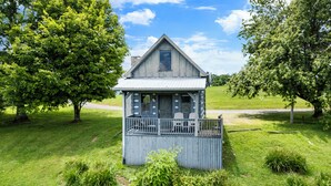 Une petite maison à un seul étage, dotée d’un porche et de deux chaises, entourée d’une pelouse verdoyante et luxuriante ainsi que d’arbres.