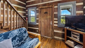 L’intérieur d’une cabane en rondins, avec un escalier en bois, un canapé à motifs floraux, un téléviseur posé sur un meuble en bois et une porte donnant sur l’extérieur.