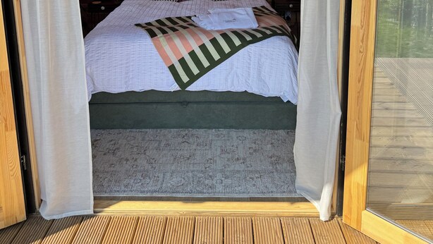 A cozy bedroom with a wooden ceiling, a bed with a green headboard, and a view of greenery outside.