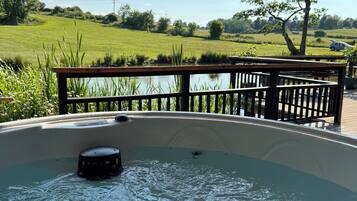 A hot tub under a wooden pergola with a view of a grassy field and a car in the distance.