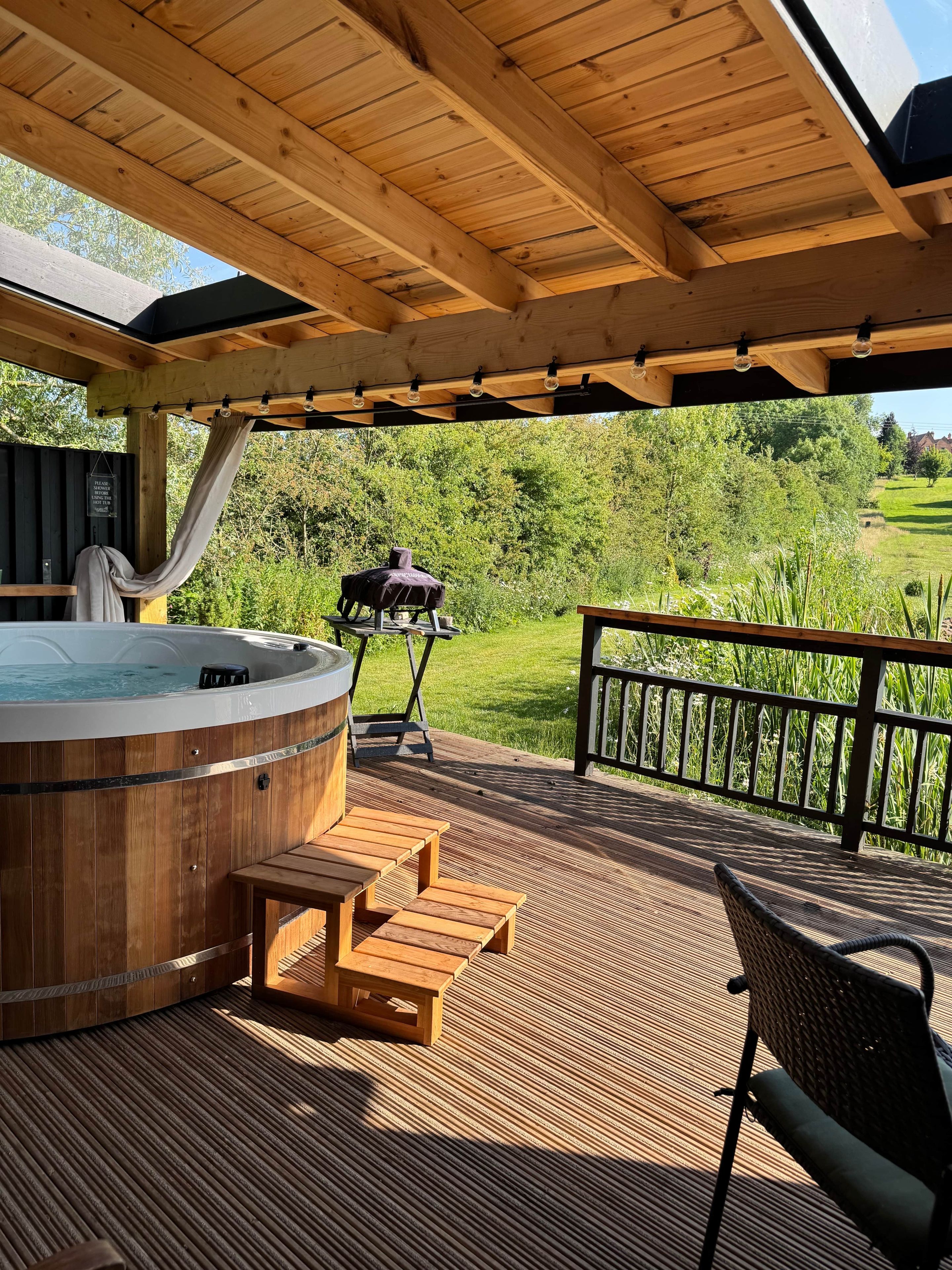 Eine Holzterrasse mit Whirlpool, Sitzgelegenheiten im Freien und Blick auf eine grüne Landschaft.