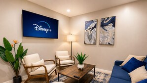 Ein Wohnzimmer mit einem Fernseher, der das Disney+-Logo zeigt, zwei Sesseln, einem Sofa, einem Couchtisch und zwei abstrakten Bildern an der Wand.