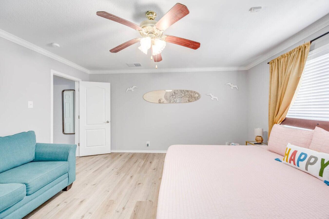 A bedroom with a ceiling fan, a bed with a 'HAPPY' pillow, a blue sofa, and a decorative wall piece.