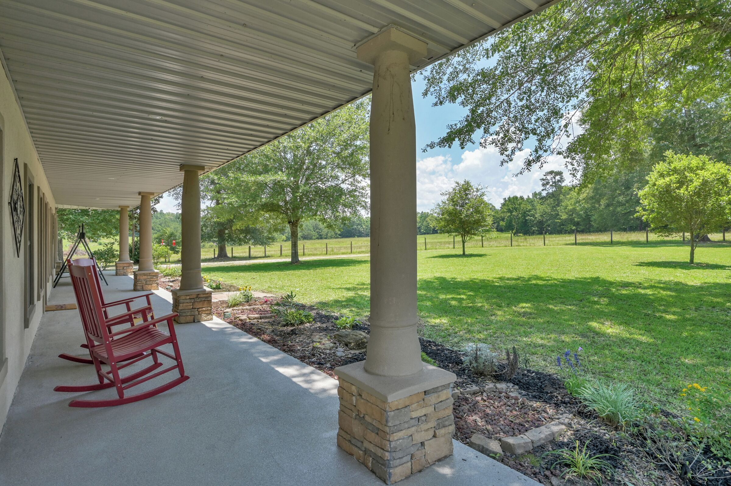 A covered porch with a stone column base, a red rocking chair, and a view of a well-maintained lawn and trees.
